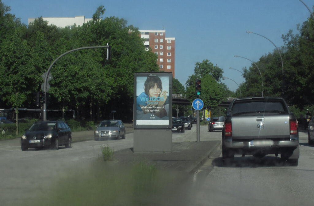 JCDecaux-Werbeglotze mit "We IQOS - Weil der Moment mir gehört", in der Mitte der Straße. Autos auf beiden Seiten.