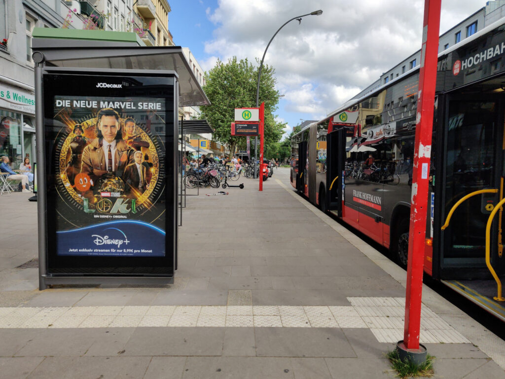 Bushaltestelle Osterstraße mit Disney Marvel Plakat, Haltestellenschild, FIMS-Anzeige und Bus.